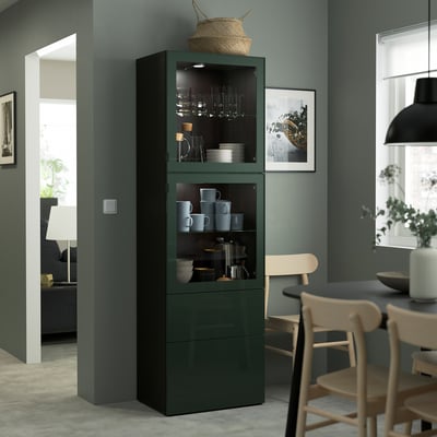 Dark green BESTÅ storage unit with glass doors dishes and mugs. Tall, stands next to dining table with wooden chairs, near a lamp and wall art.