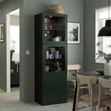 Dark green BESTÅ display cabinet, tall, with glass doors, holding cups & bowls, next to wooden chairs by dining table.
