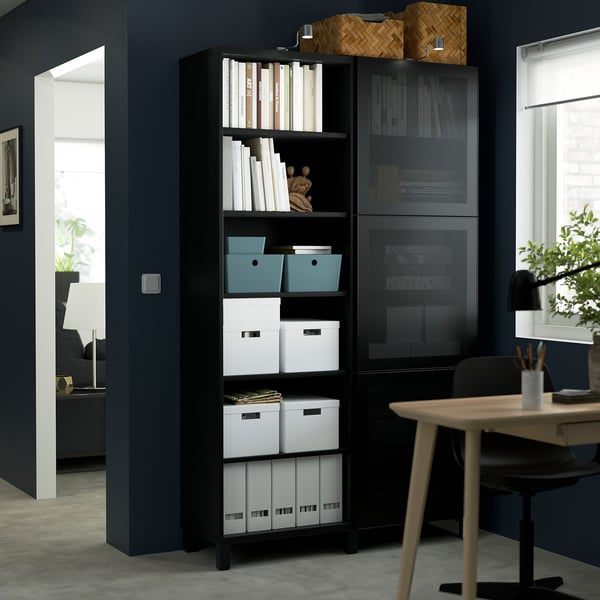 Black BESTÅ storage with shelves & glass doors, holding books, boxes, decor, in modern room with dark blue walls, next to desk & chair.