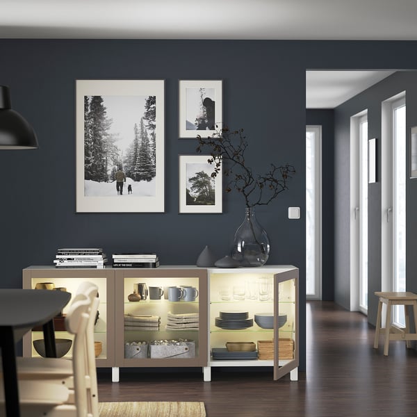 Modern living room with gray walls, illuminated BESTÅ sideboard showing dishes and a vase, nature photos above, right hallway.