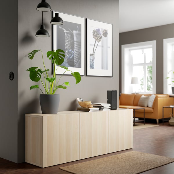 Wooden BESTÅ sideboard with books, speaker, plant. Cozy living room with sofas and window.