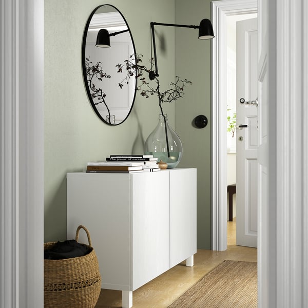 White BESTÅ sideboard with adjustable shelves and doors; light fixture, mirror, books, vase with branches, woven basket.