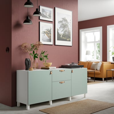 Modern living room with green BESTÅ sideboard, vases, bowl. Red wall, framed pictures, black lamps. Orange sofa by window.