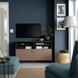 Modern living room with BESTÅ tv bench in pinkish-brown/black. Tv, shelves, vase, lamp, basket, blue armchairs, & personal photos.