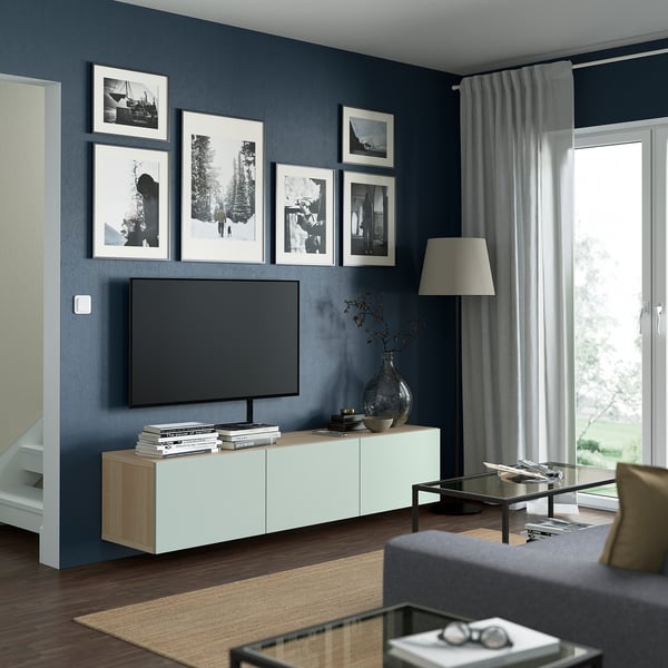 Modern living room with BESTÅ tv bench, flat screen tv, photos, grey walls, glass coffee table, sofa, floor lamp, and large window with curtains.