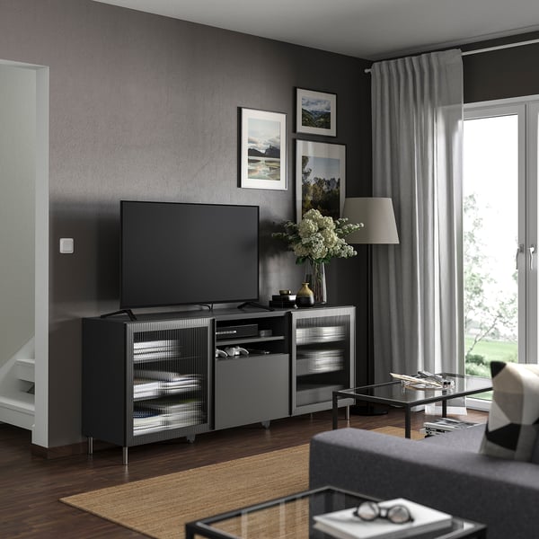 Modern living room with BESTÅ tv bench in gray, large flat-screen tv, shelf with books, lamp, vase with flowers, curtains, and large window.