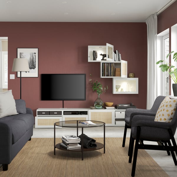 Living room with red wall, grey sofa, glass coffee table, tv, white storage with shelves, terra cotta rug, and black easy chair.