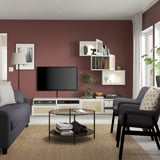 Living room with red wall, grey sofa, glass coffee table, tv, white storage with shelves, terra cotta rug, and black easy chair.