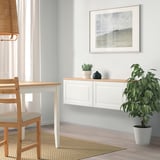 Minimalist dining room with BESTÅ sideboard, table, wicker chair, plant, and wall frame.