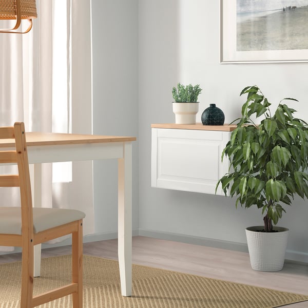 A minimalist dining room with BESTÅ sideboard, plant, and light wooden chairs around white wall-mounted table.