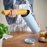 Person holding light blue BETTHAJ device with spaghetti inside, showing kitchen scene.