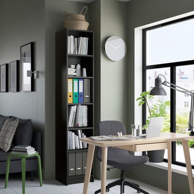 Black BILLY bookcase packed with books, beside window and chair. Tall, functional design with particleboard.