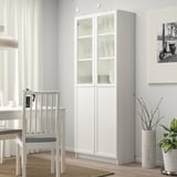 High white BILLY cabinet with glass doors, holding plates, next to small plant, gray chair by dining table.