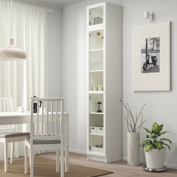 White BILLY bookcase with glass doors. Beside it, a dining set, framed picture, and plant. White curtains.