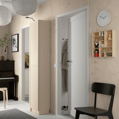Modern entryway with light-colored wooden walls, an open closet with hangers, a tall BILLY bookcase, and a black chair.