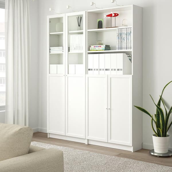 White BILLY bookcase with glass doors, holding books and decor, next to a beige chair and a large potted plant.