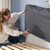 Woman fixing a gray upholstered bed frame with soft padding, showing comfort and easy maintenance.