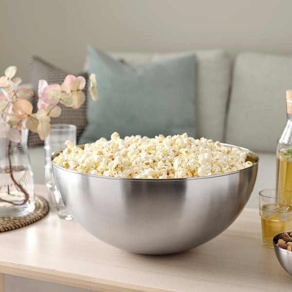 Large metal bowl filled with popcorn on a table, surrounded by glasses, a bottle, and a vase with flowers.