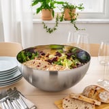 Large stainless-steel bowl filled with salad on a dining table, surrounded by glasses, plates, and cutlery, showcasing its use for serving food.