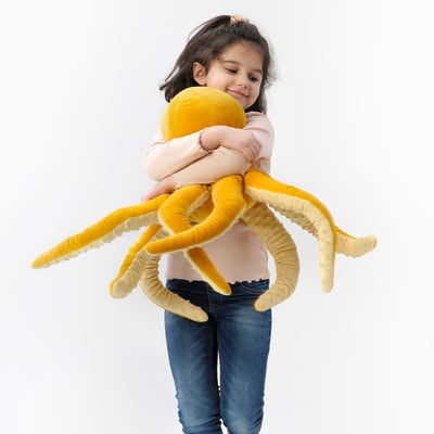 In a child hugs a large yellow plush octopus toy. The toy has soft, long tentacles and is part of the BLÅVINGAD collection.