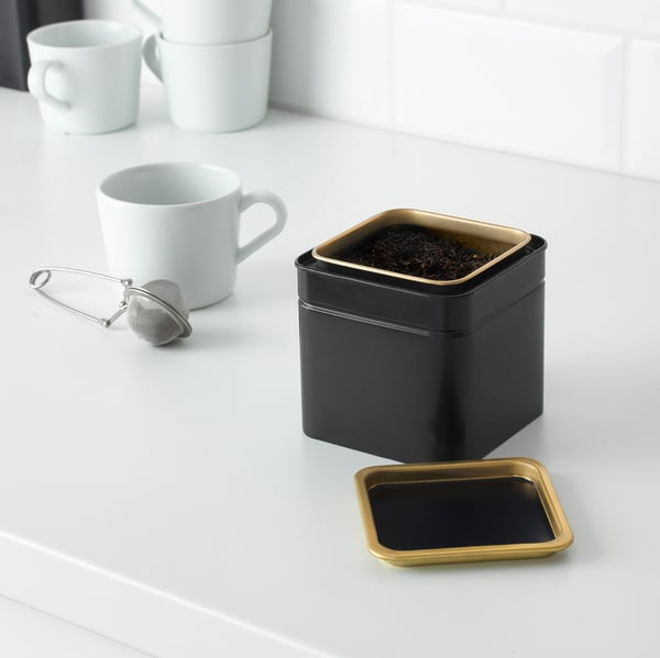 Black metal tea tin with gold lid sits on white counter, guarding tea leaves. Two white mugs and a tea infuser nearby, ready for brewing.