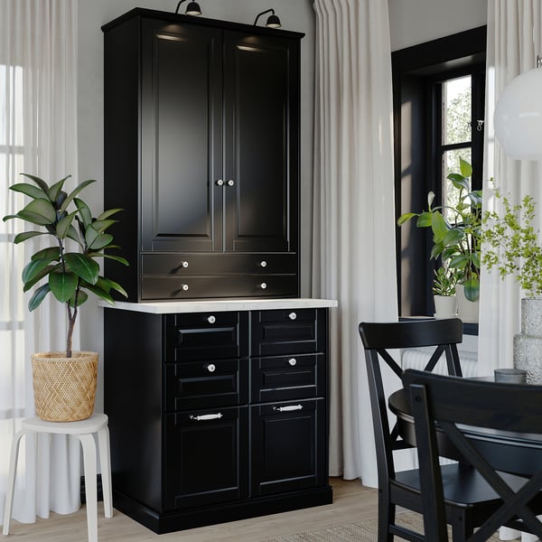 Black BODBYN cabinet with drawers and doors, lacquered fiberboard, classic beveled panel design, near window with plants.