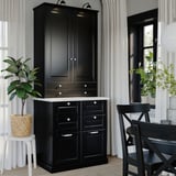 Black BODBYN cabinet with drawers and doors, lacquered fiberboard, classic beveled panel design, near window with plants.