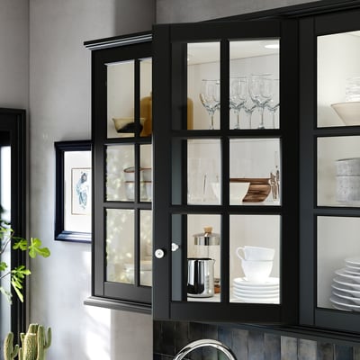 Black BODBYN glass cabinets display glassware and dishes, framed with beveled glass, mounted on a gray wall.