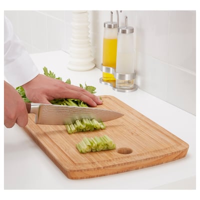 Person chops green vegetables on wooden cutting board with sharp knife. Bamboo board on white counter, stainless dispensers in background.