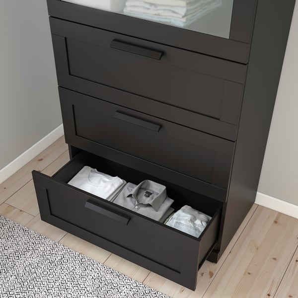 Black chest of drawers with frosted glass top, one drawer open showing folded clothes. Wall-anchored, smooth-running drawers.