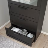 Black chest of drawers with frosted glass top, one drawer open showing folded clothes. Wall-anchored, smooth-running drawers.