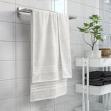 BROKGLIM white towel hangs neatly, bordered. Surrounded by gray towels, plant, and tiles. Stainless hanger.