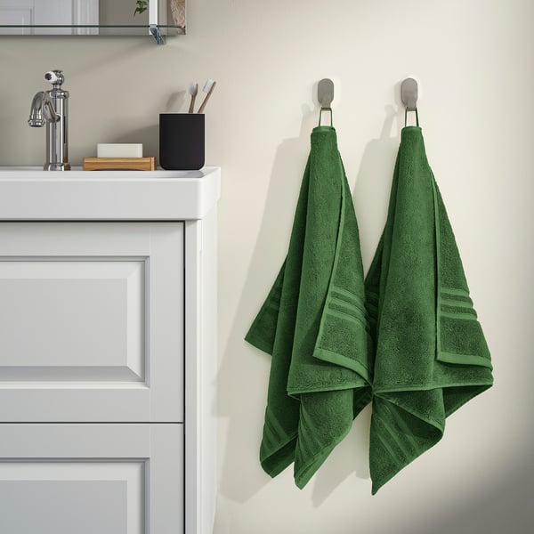 BROKGLIM organic towels in bathroom. Dark green, 100% cotton, folded on silver hooks beside sink. Soft, absorbent, with woven border.