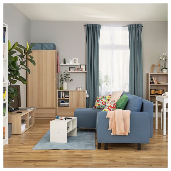 Modern living room with wooden floors, blue sofa-bed, white storage, large window, and tall wardrobe.
