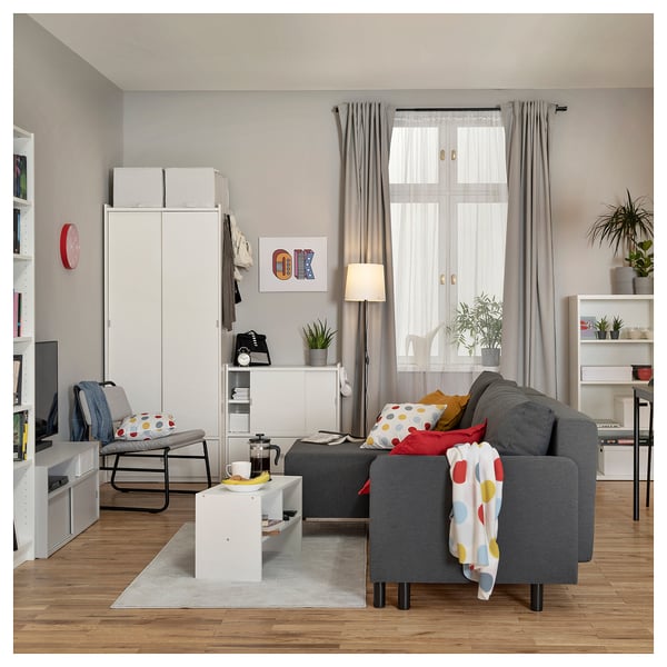 Modern living room with grey l-shaped sofa-bed, white storage units, colorful pillows, plants, and wooden floor.