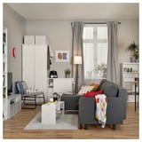 Modern living room with grey l-shaped sofa-bed, white storage units, colorful pillows, plants, and wooden floor.