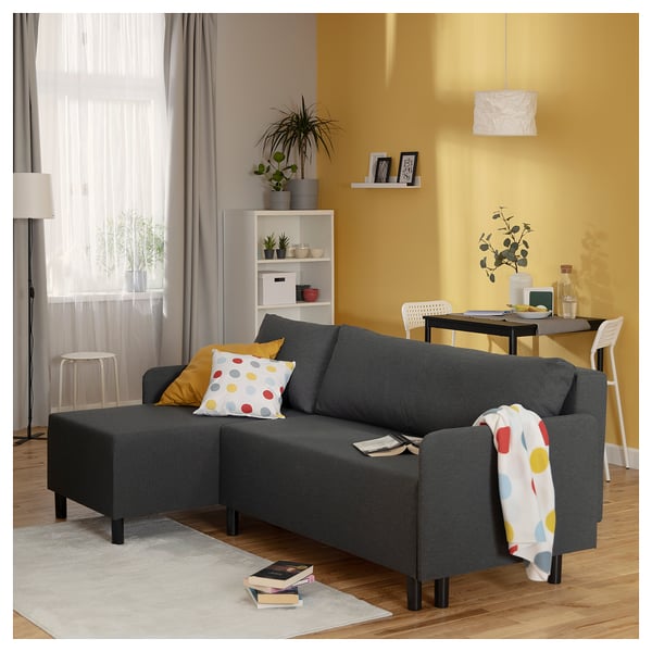 Living room with gray BRUKSVARA l-shaped sofa-bed, colorful pillows, books, plants, and a dining area with yellow walls.