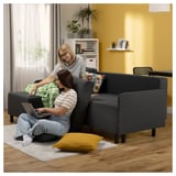 Two women relax on a gray BRUKSVARA l-shaped sofa-bed with colorful pillows in a cozy yellow room.