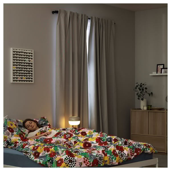 A cozy bedroom with dark gray curtains, a colorful floral-patterned bed, a lampshade, and wall art; a person sleeps comfortably.