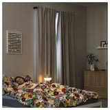 A cozy bedroom with dark gray curtains, a colorful floral-patterned bed, a lampshade, and wall art; a person sleeps comfortably.