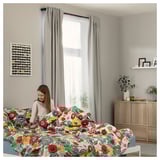 Woman on floral bed in cozy room. Bright patterns, plant, frames.