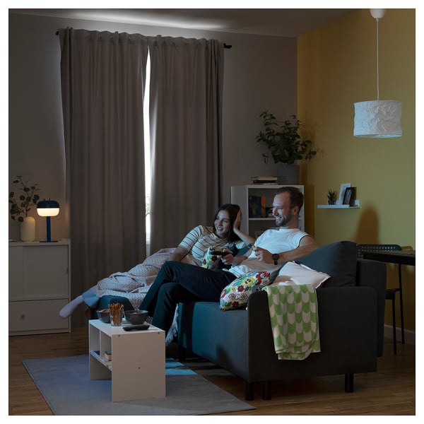 Couple playing video games on sofa in cozy living room with curtains and lamp.