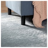 A light blue rug with black legs supporting a gray upholstered piece, accented with a fringe throw.