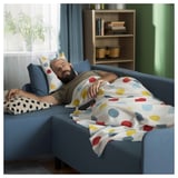 A man naps on a blue BRUKSVARA sofa, covered with a white throw blanket featuring colorful spots. The room has wooden shelves with books and plants.