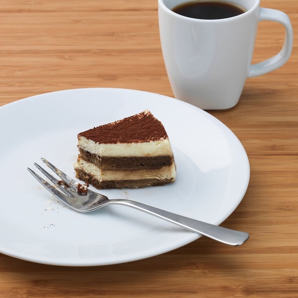A slice of layered dessert with a fork on a white plate, beside a coffee cup.