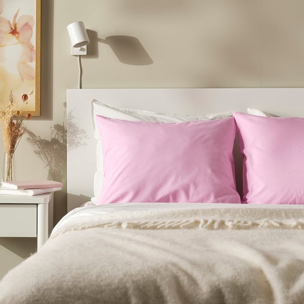Bed with large, soft pink DVALA pillowcases; white headboard, cosy beige blanket, wall lamp, and floral artwork.