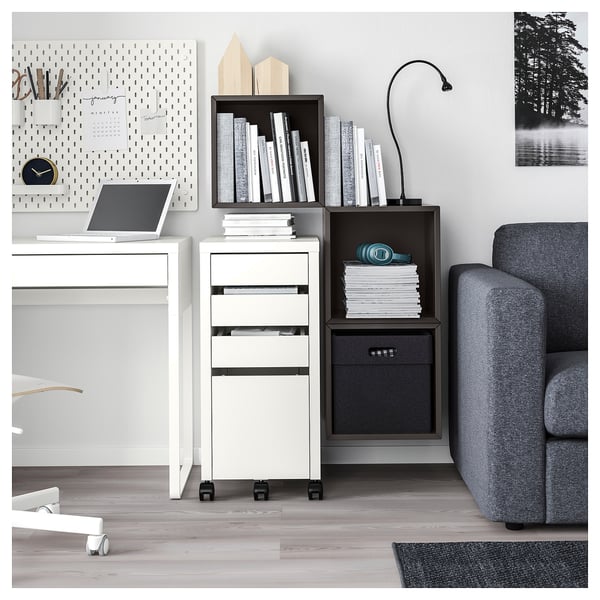 Modern office setup with white EKET storage units, containing books and files. Minimalist desks and gray upholstered couch.