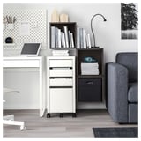 Modern office setup with white EKET storage units, containing books and files. Minimalist desks and gray upholstered couch.