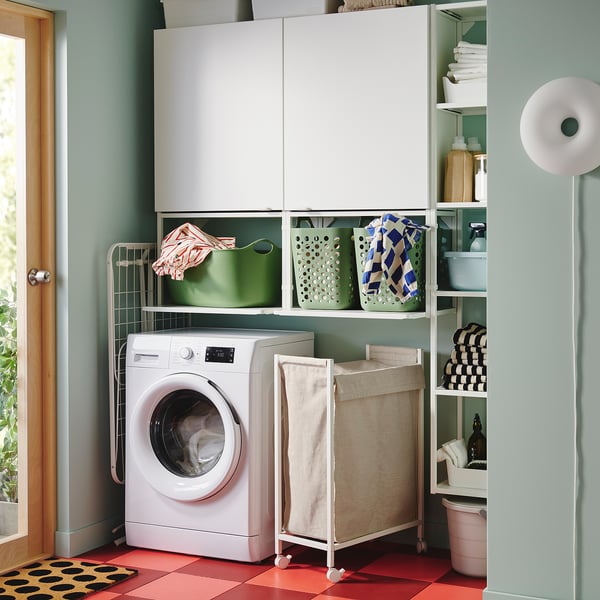 ENHET white laundry setup: washing machine, shelves with towels, cleaning supplies, cabinet, rolling hamper, chequered floor, green walls.