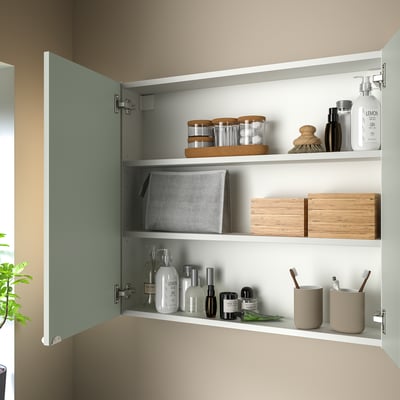 Open wall cabinet displaying toiletries and storage boxes. Pale gray-green front and three shelves.